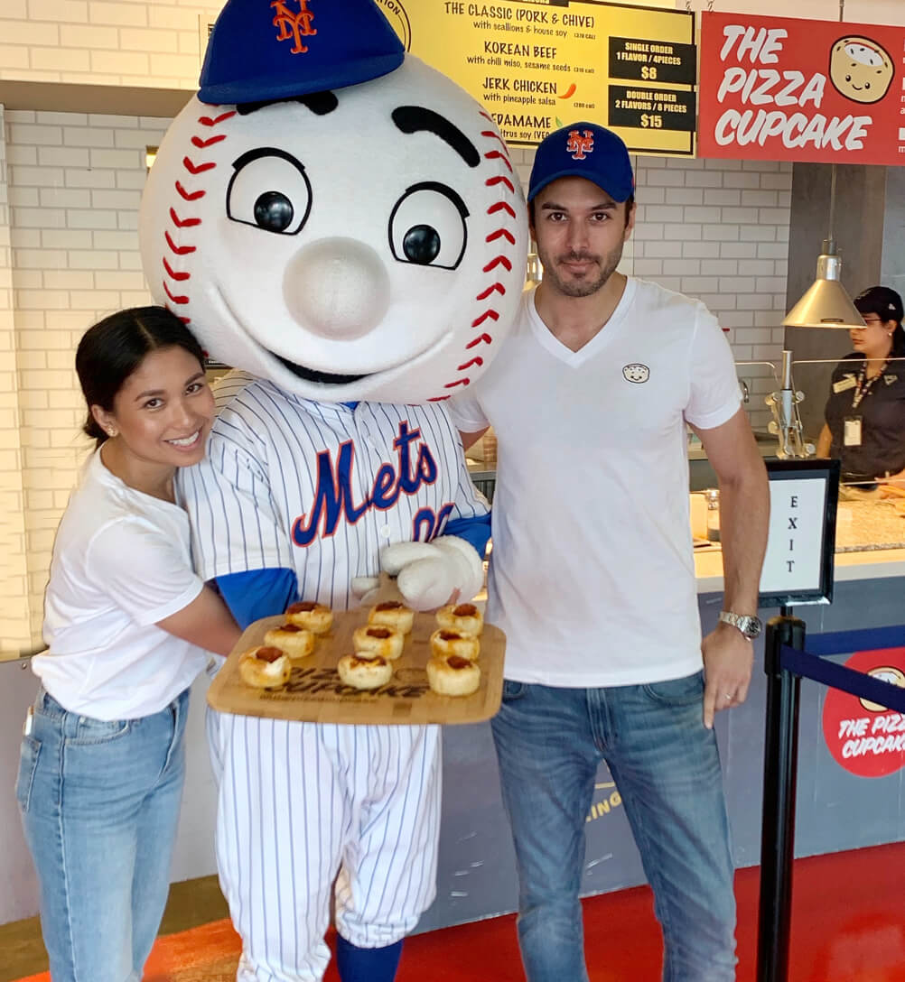 A man and woman smile with a New York Mets mascot, holding a pizza cupcake, posing for the camera.