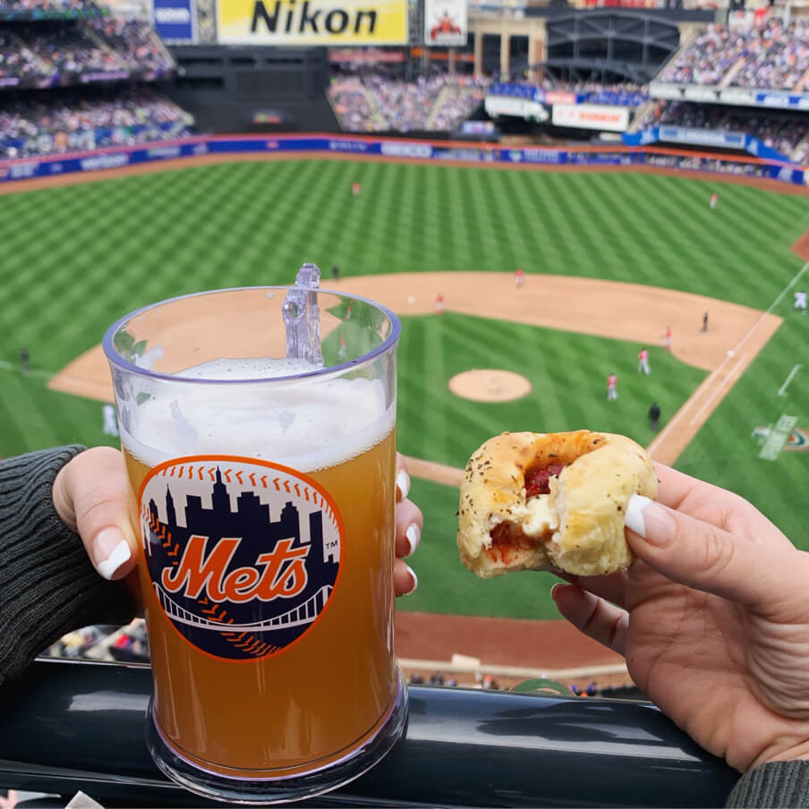 A person enjoying a beer and a pizza cupcake at a lively baseball game.
