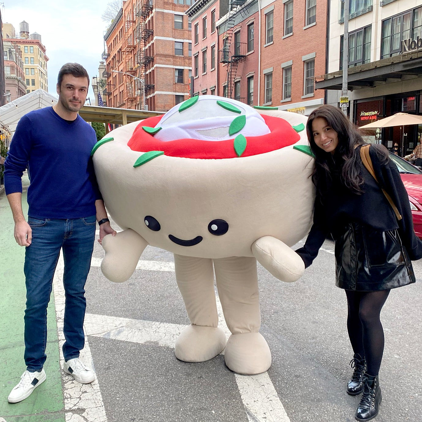 Owners Andrea and Michelle stand on a street next to a large, plush pizza cupcake mascot