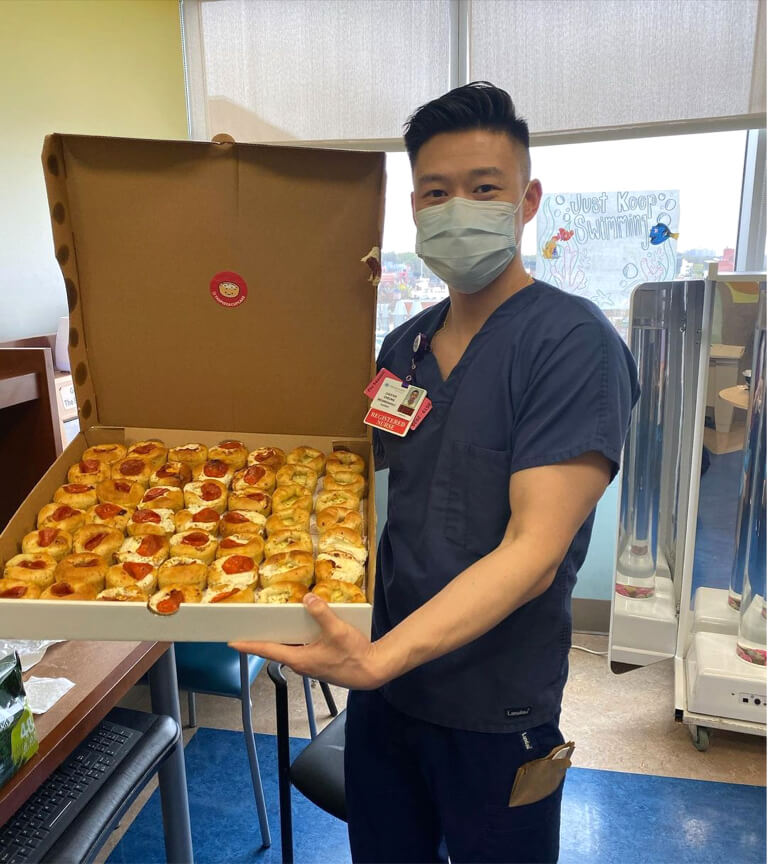 A man in scrubs holds a pizza box, standing next to a cupcake shaped like a slice of pizza.